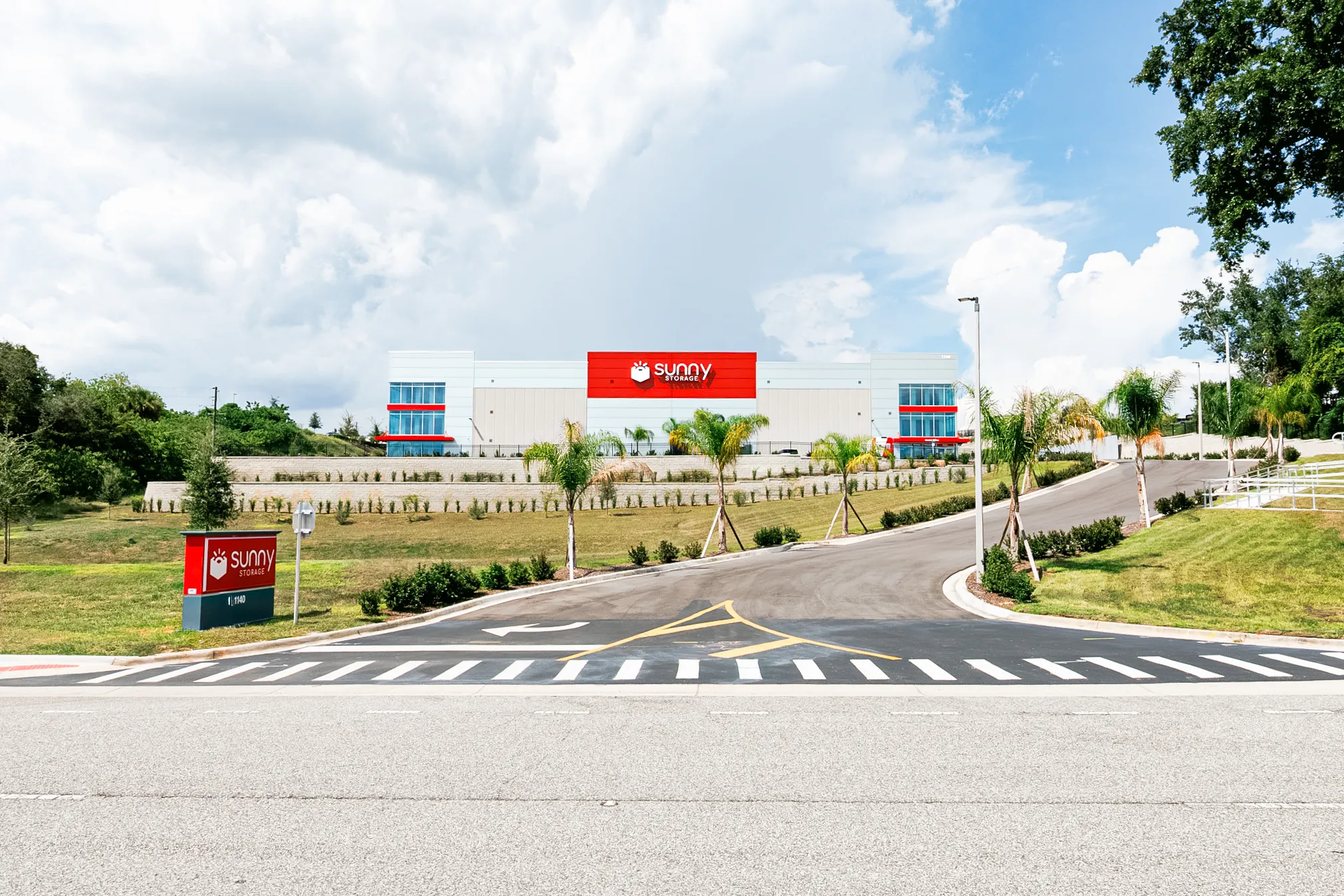 Sunny Storage self-storage building on Hooks St. in Clermont, Florida, seen from the entrance drive, with the red Sunny Storage sign on a white and grey facade, palm trees and a cloudy blue sky.